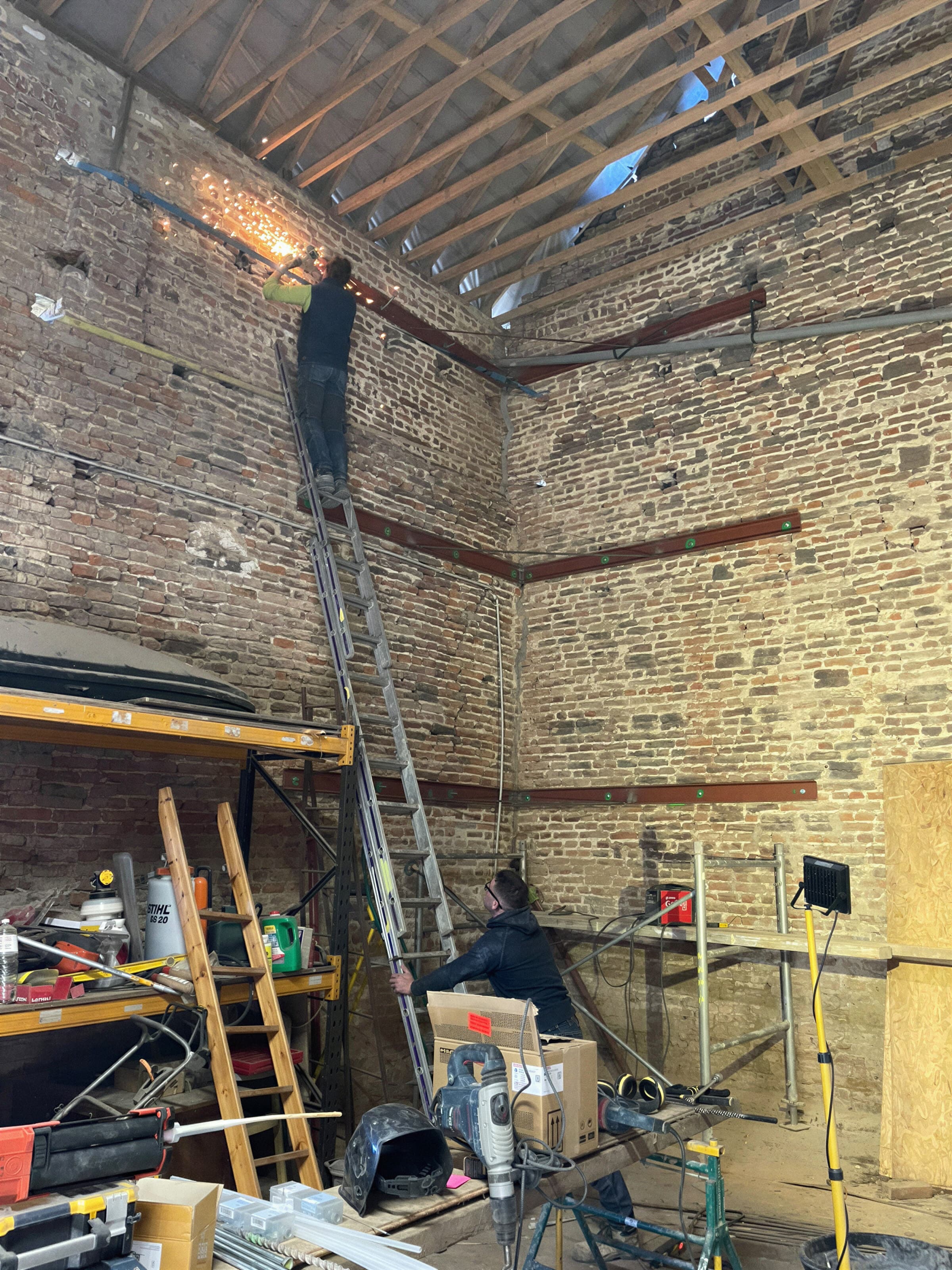 Artisans au travail sur un mur en briques et moellons, échafaudage et échelle dans une grange en rénovation